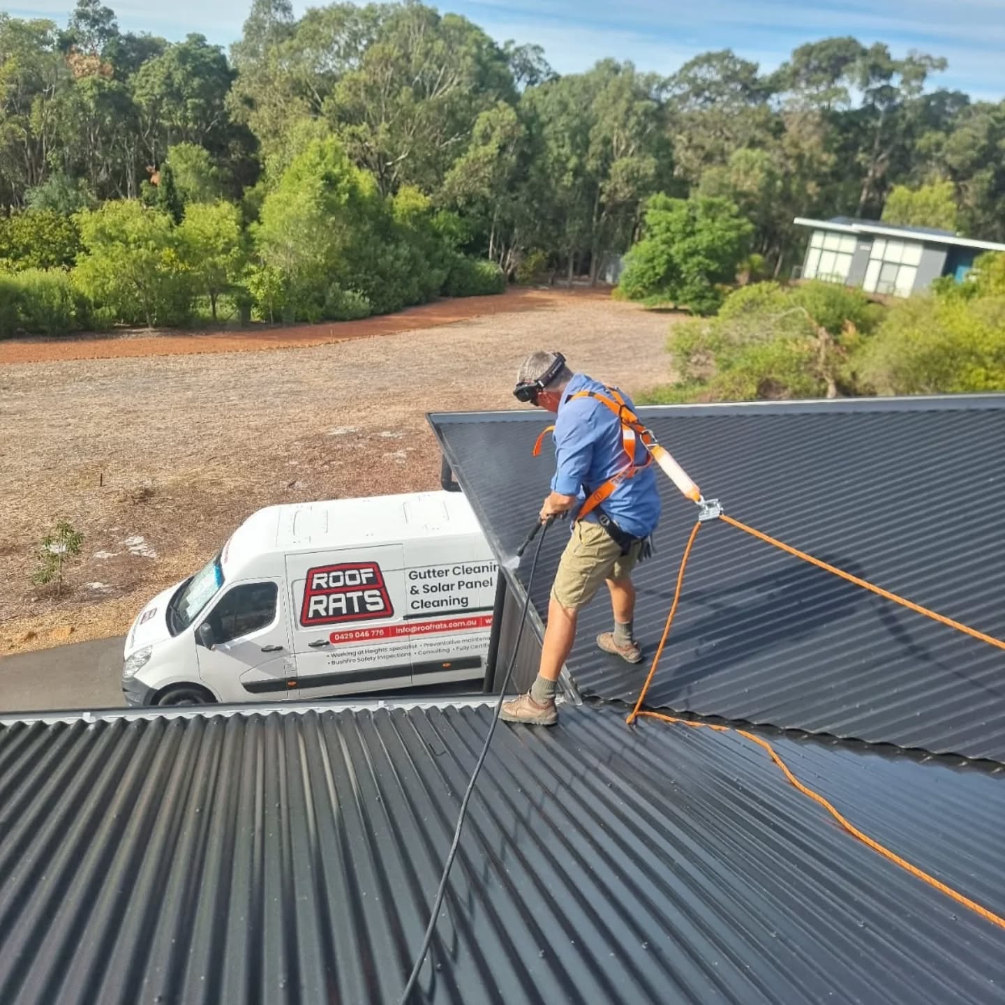 Worker in a harness on a dark metal roof with the Roof Rats van parked below